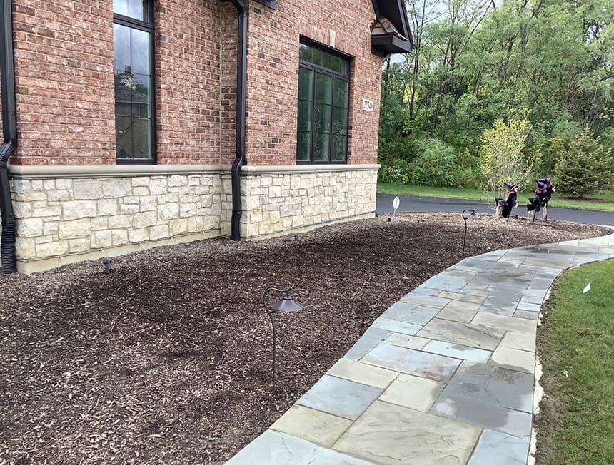 Before renovation view of a foundation planting area in Kildeer, Illinois, showing bare mulch beds alongside a stone walkway and brick home.