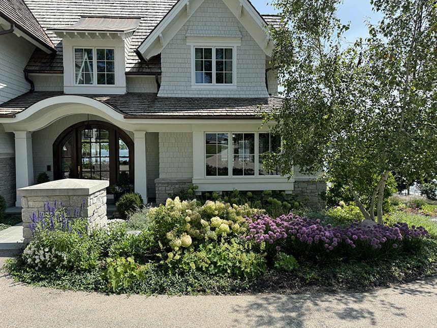 Completed front landscape design at a custom lakeside home in Williams Bay, Wisconsin, featuring layered perennial plantings, natural stone edging, and a cottage-style exterior.